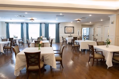 A spacious dining room with multiple tables covered in white tablecloths, each surrounded by wooden chairs with cushioned seats. Small flower arrangements are placed on each table. The room has wooden flooring, large windows with blue curtains, and soft ceiling lighting.