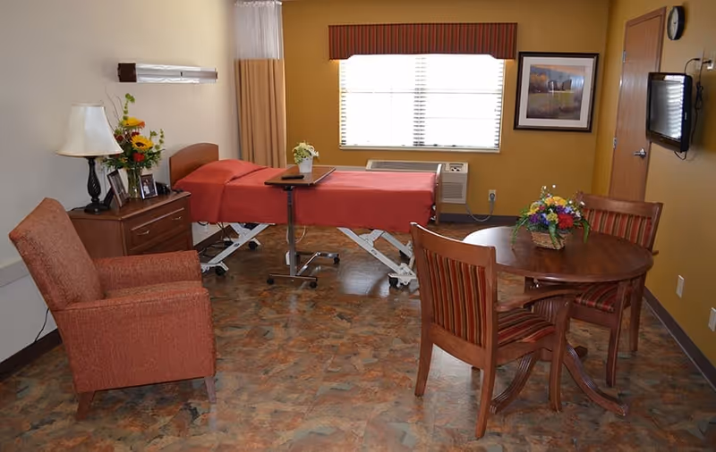 A well-lit room with a medical bed covered in a red blanket, a wooden side table with a lamp and flower vase, a comfortable armchair, a round wooden table with three striped chairs, and a window with blinds and a valance. The room has a warm color scheme with yellow walls and a patterned floor.