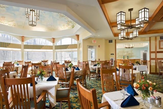 A bright dining room with multiple set tables and wooden chairs, floral centerpieces, and large windows.