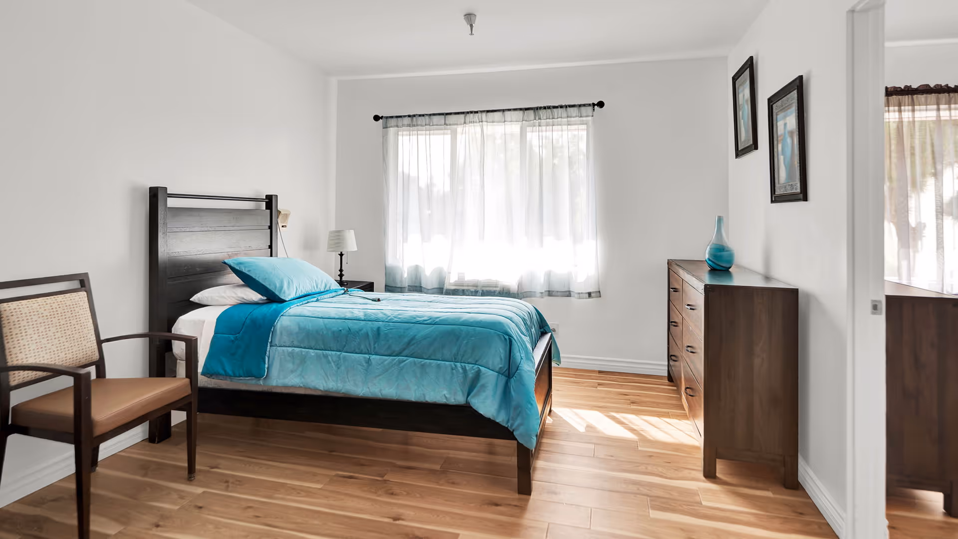 A bright bedroom with a wooden bed frame and a turquoise comforter and pillow. There is a wooden chair with a cushioned seat to the left of the bed, a small bedside table with a lamp, and a wooden dresser with a blue vase on top. Two framed pictures hang on the wall above the dresser. Light filters through a window with sheer white curtains.