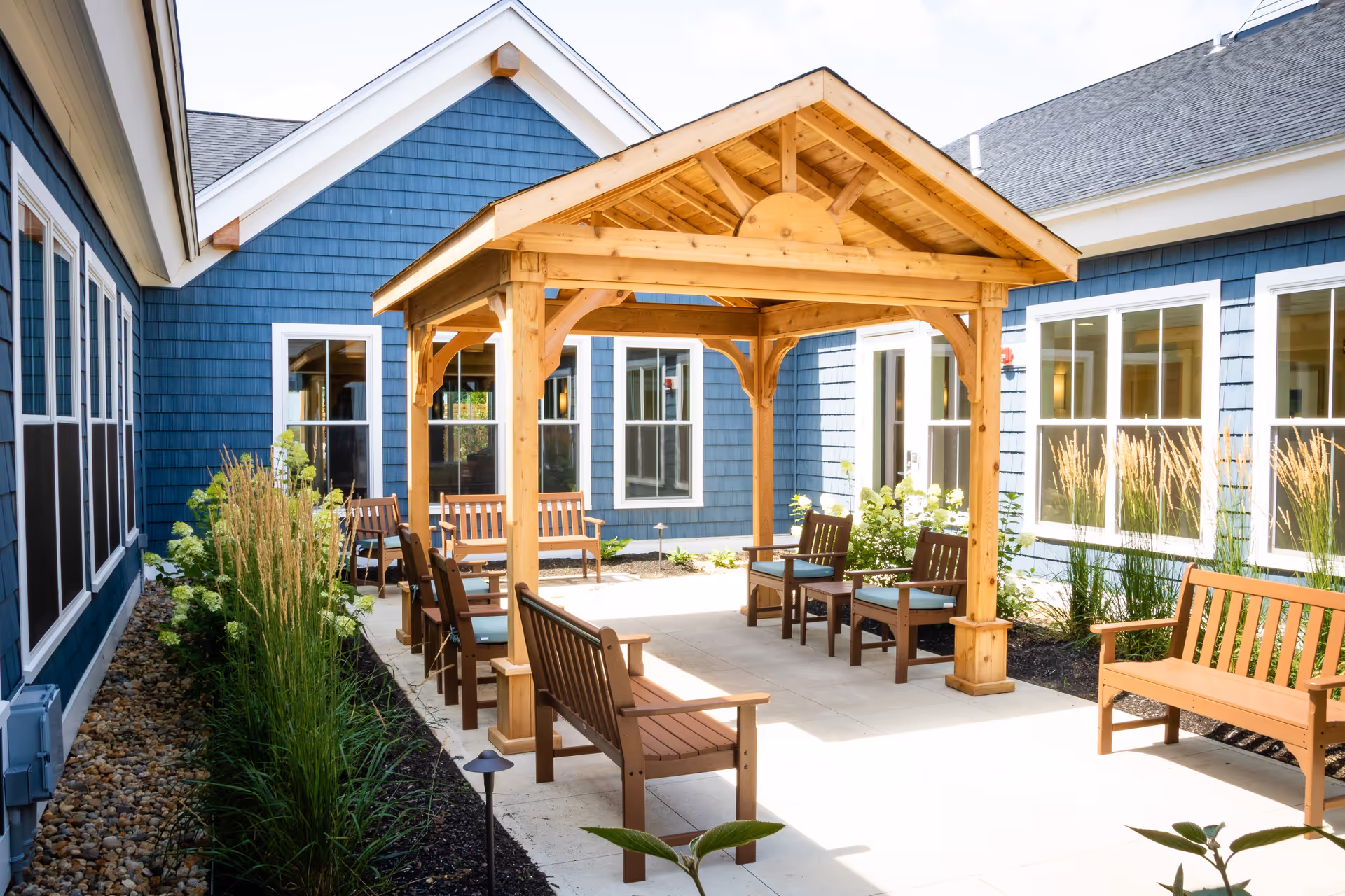 Sunny courtyard with a wooden pergola and multiple benches and chairs between blue-sided building facades.