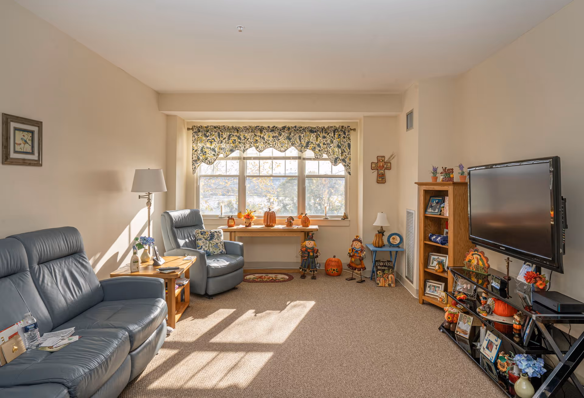 A cozy living room with two blue leather recliners, a wooden side table with a lamp and flowers, a large window with floral valance, and various autumn-themed decorations including pumpkins and scarecrows. There is a wooden bookshelf and a wall-mounted flat-screen TV on the right side.