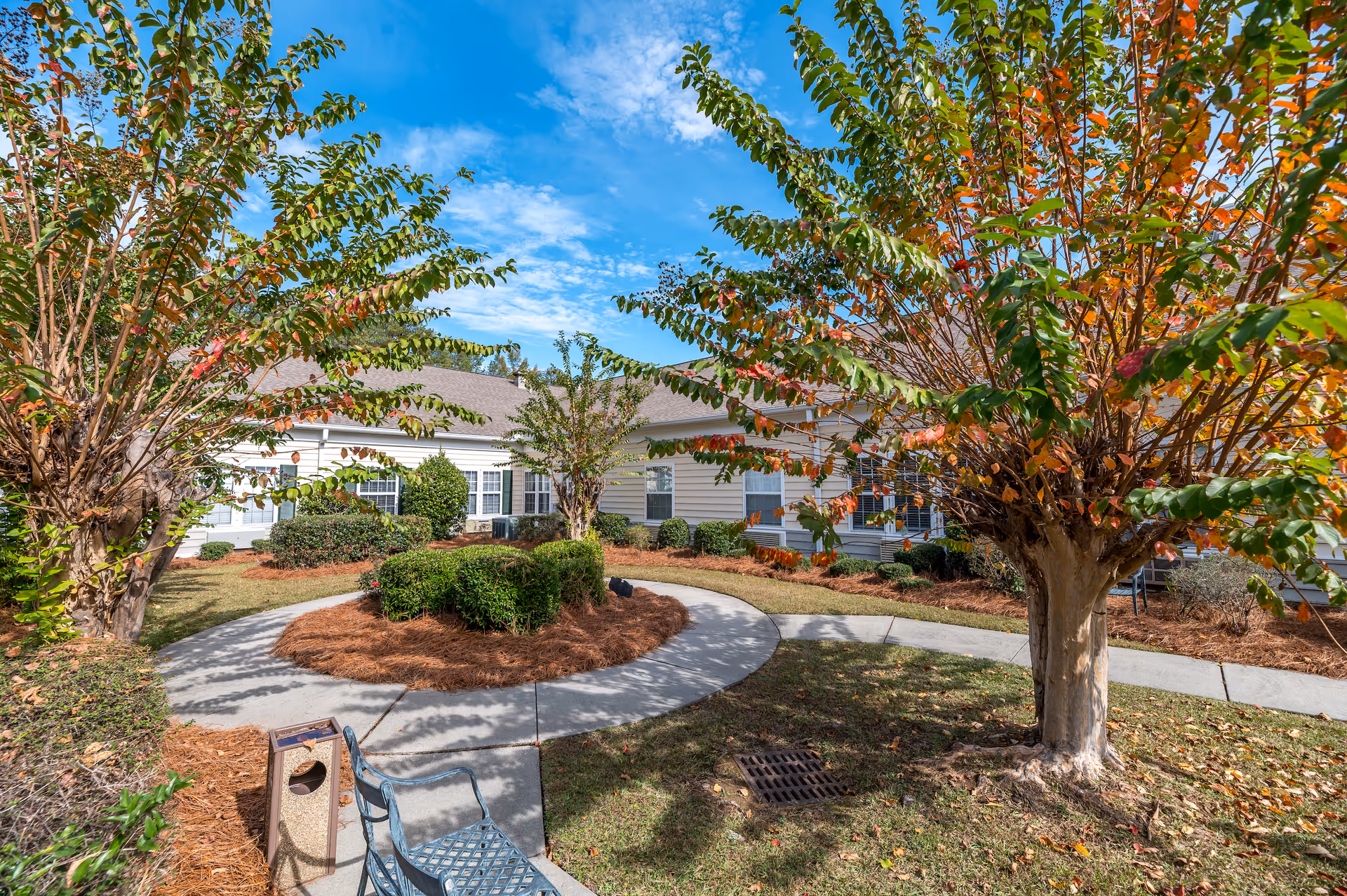 Courtyard with a circular walkway, trees, shrubs, a bench, and a single-story senior living building in the background under a blue sky.