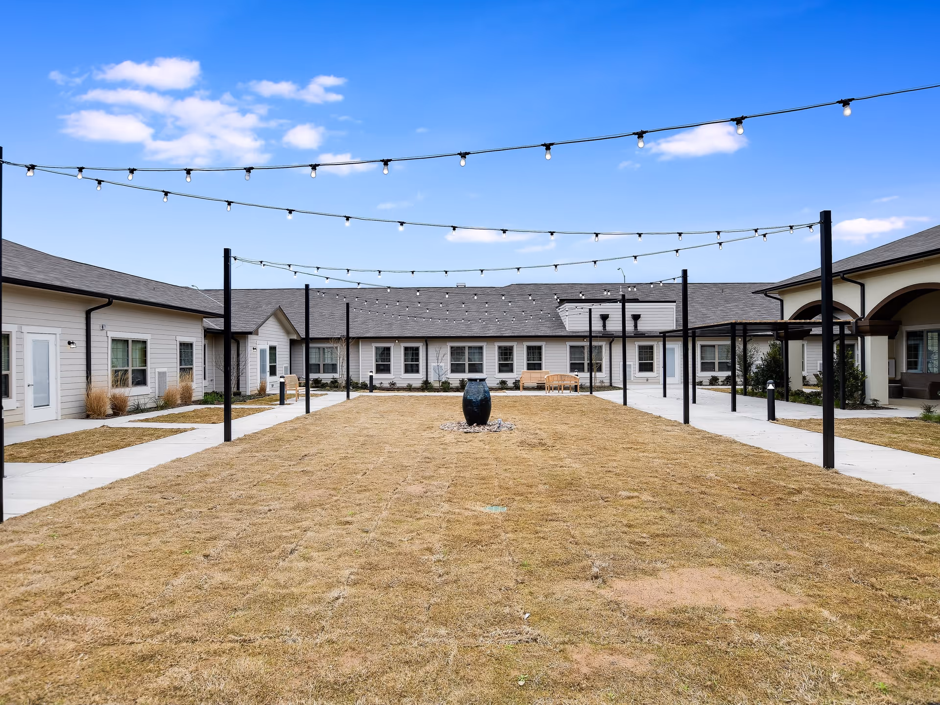 Outdoor courtyard area at The Grandview of Chisholm Trail featuring a central grassy lawn with a decorative black urn, surrounded by a paved walkway and single-story buildings. String lights are hung overhead across the courtyard under a blue sky with some clouds.