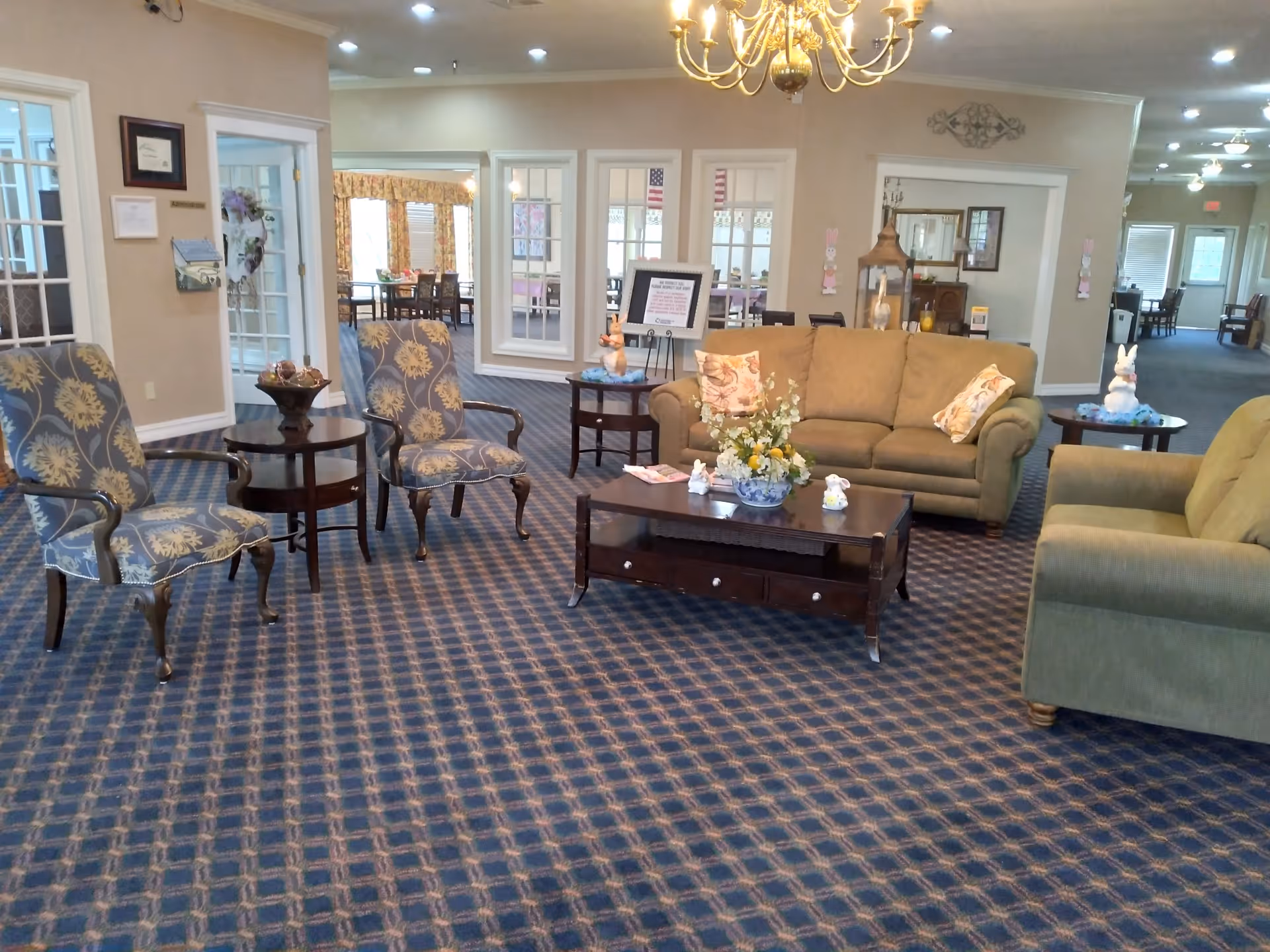 A spacious assisted-living common lounge with sofas, patterned armchairs, and coffee tables on a patterned carpet.