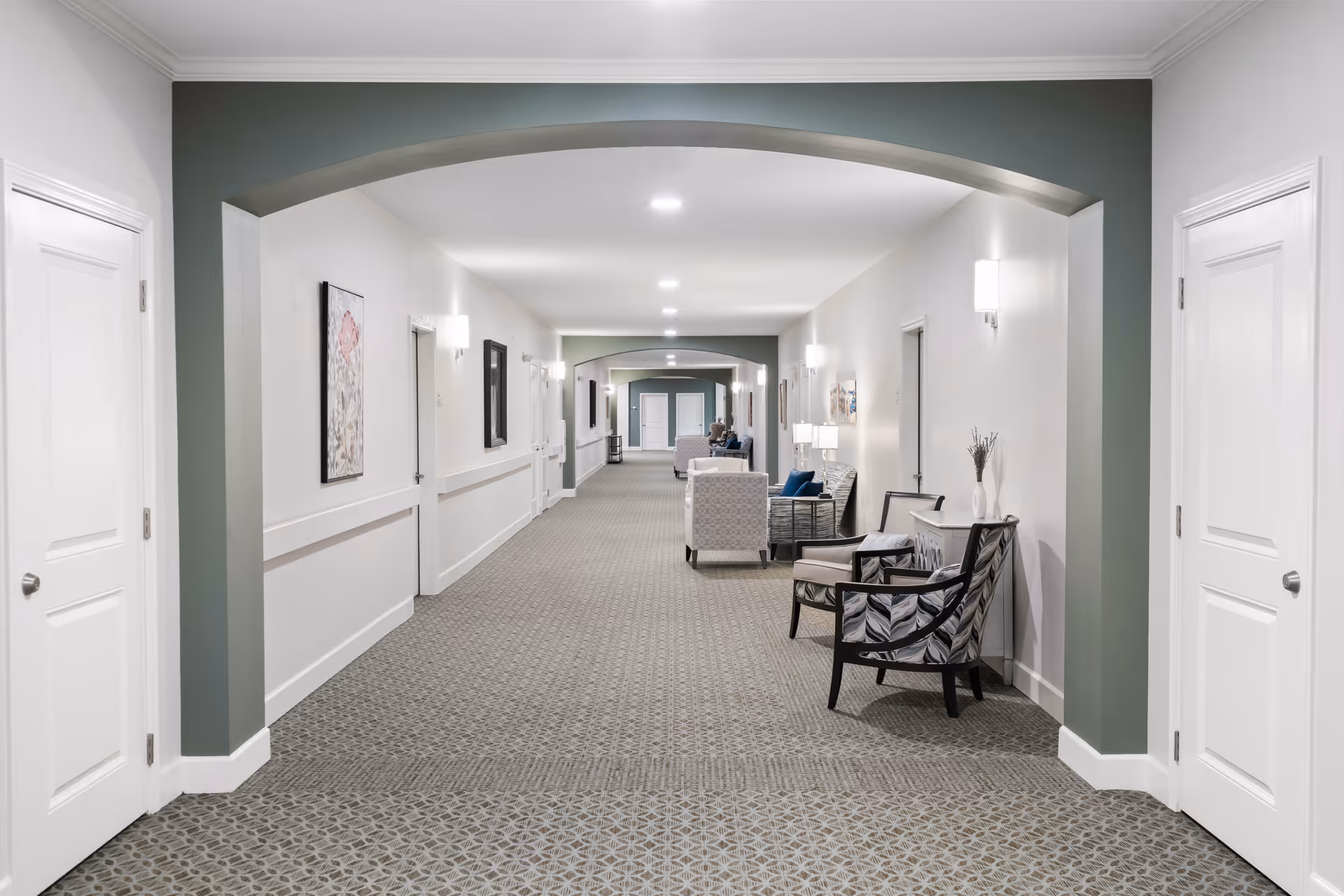 A long, well-lit hallway in a senior living facility with white walls and patterned carpet. The hallway features multiple white doors on both sides, wall-mounted lights, framed artwork, and several seating areas with armchairs and small tables. The ceiling has recessed lighting and there are archways along the corridor.