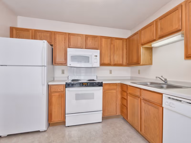 A clean kitchen with wooden cabinets, a white refrigerator, a white microwave above a white stove, a double sink, and a white dishwasher. The countertops are light-colored and the floor has a light tile pattern.