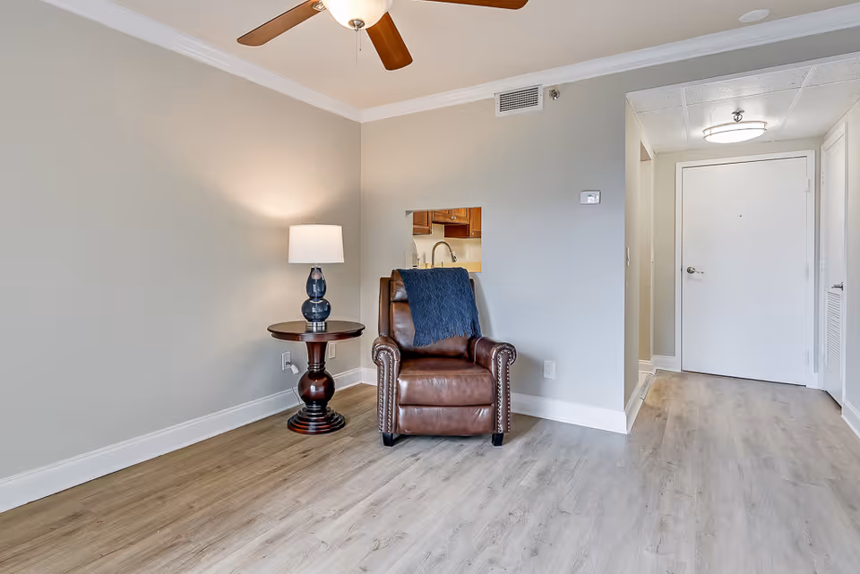 Leather recliner beside a round side table with a lamp in a bright living area with an entry door and ceiling fan.