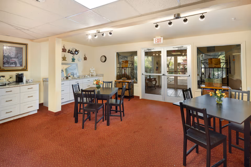 A cozy dining area in a senior living community with two black tables, each surrounded by four black chairs. The room has a red carpet with a dotted pattern, cream-colored walls, and a white ceiling with recessed lighting. There is a countertop with cabinets along one wall, decorated with coffee-themed wall art and a clock. Double glass doors lead outside, allowing natural light to enter the room.
