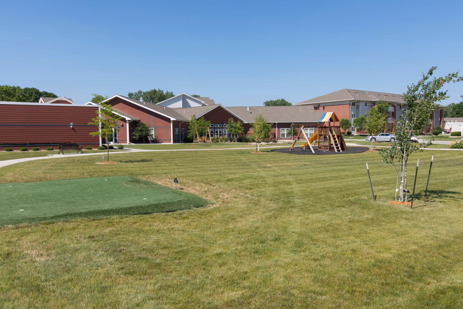 Outdoor view of Sunrise Retirement Community showing a large grassy area with a small putting green, a playground with a slide and swings, several young trees, and red buildings in the background under a clear blue sky.