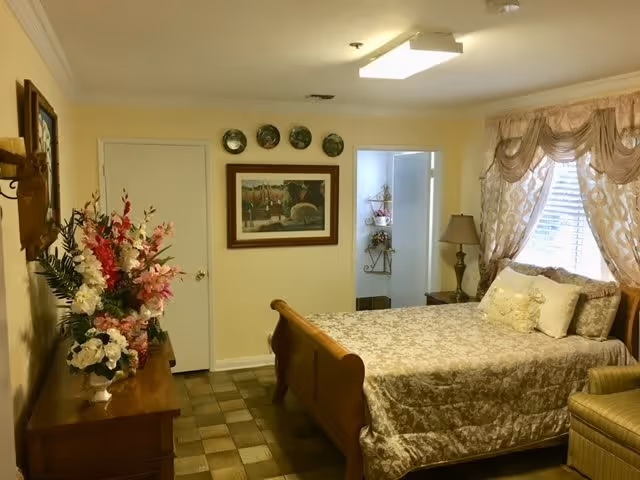 Sunlit bedroom with a wooden sleigh bed, floral bedding, dresser topped with flower arrangements, a bedside lamp, and framed wall art.