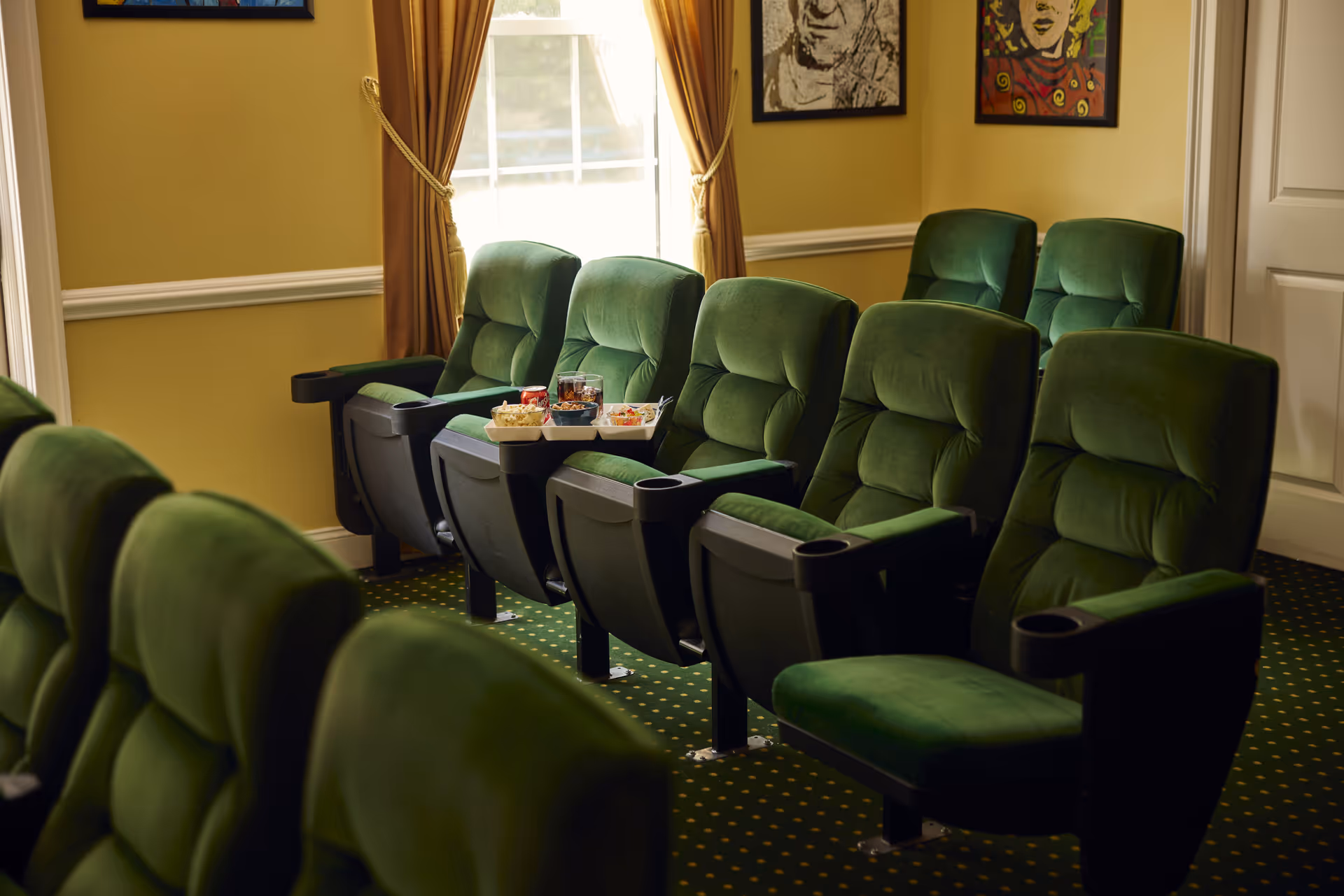 A small theater room with two rows of green cushioned reclining seats. A window with brown curtains is in the background, and there are framed artworks on the yellow walls. Snacks and drinks are placed on the armrest of one of the seats.