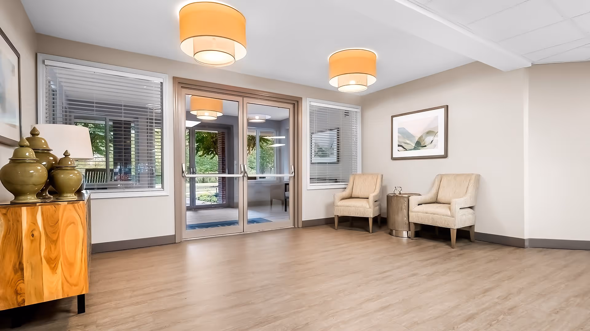 A bright and clean sitting area in a senior living facility with two beige armchairs separated by a small round wooden table, a wooden cabinet with decorative green vases and a lamp, large windows with blinds, and double glass doors leading outside. The room has light wood flooring, neutral-colored walls, and two ceiling lights with beige lampshades.