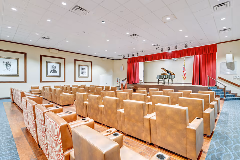 A small auditorium with rows of tan theater seats facing a stage with a grand piano and red curtains.