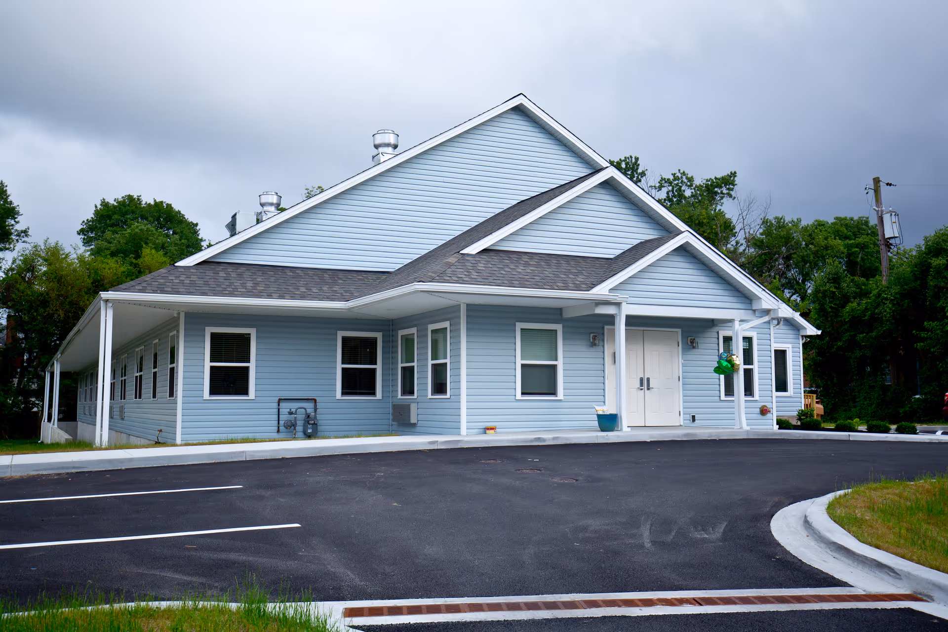 Exterior view of a light blue single-story building with white trim, a gabled roof, and a covered entrance with double doors. The building is surrounded by a paved parking lot and greenery, under a cloudy sky.