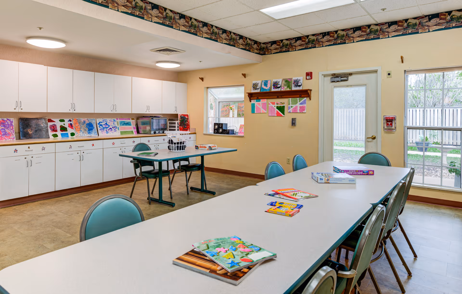 A bright activity room with a long white table surrounded by green chairs. On the table are various coloring books and art supplies. The room has white cabinets along one wall with colorful artwork displayed above them. There is a smaller table with chairs and a laundry basket, a microwave on the counter, and a door and windows letting in natural light.