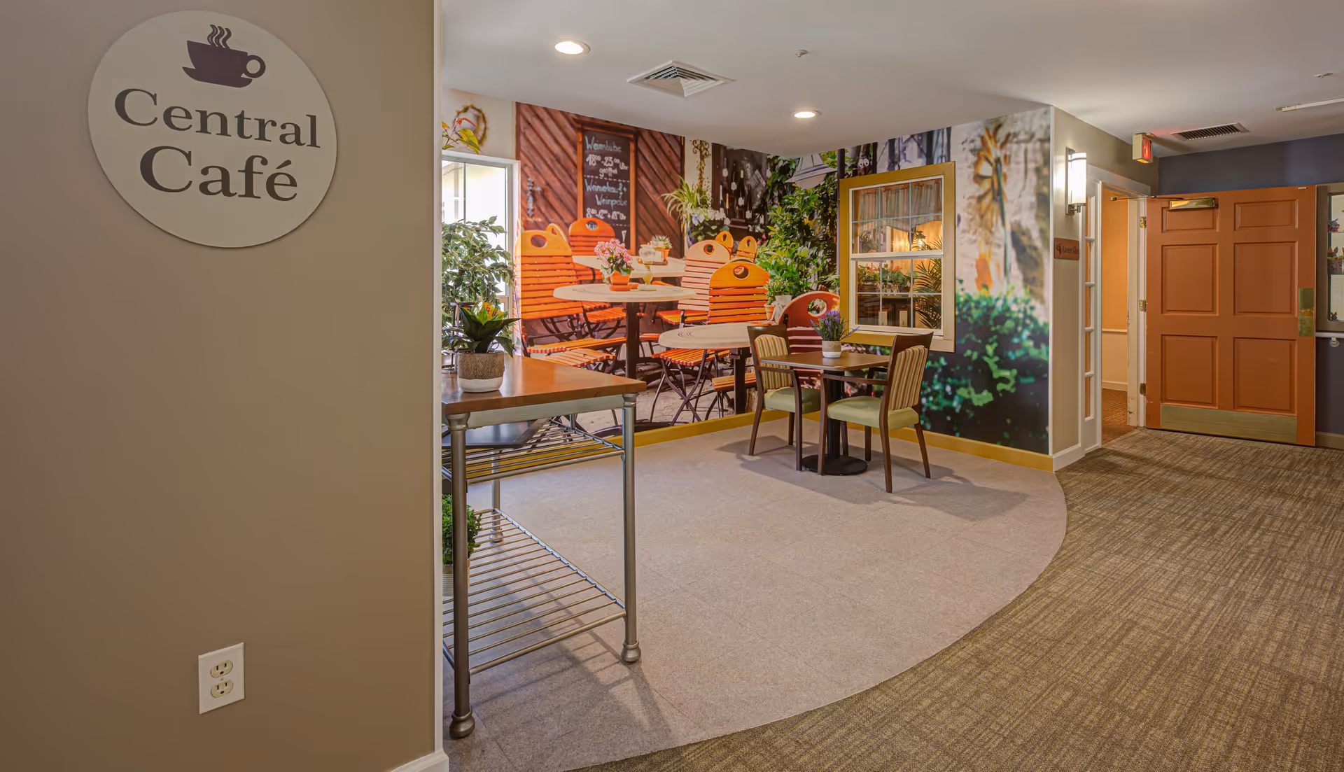 Interior view of a cozy café area named Central Café with a small table and two chairs, a metal cart with plants, and a large wall mural depicting an outdoor café scene with orange chairs and tables surrounded by greenery.