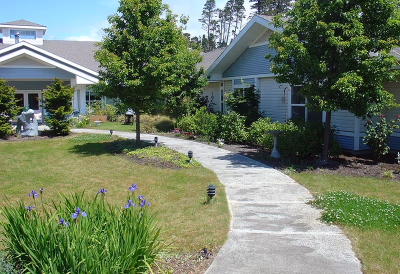 A curved concrete pathway leading through a well-maintained garden with green grass, flowering plants, and small trees, surrounded by single-story buildings with light-colored siding and pitched roofs under a clear blue sky.