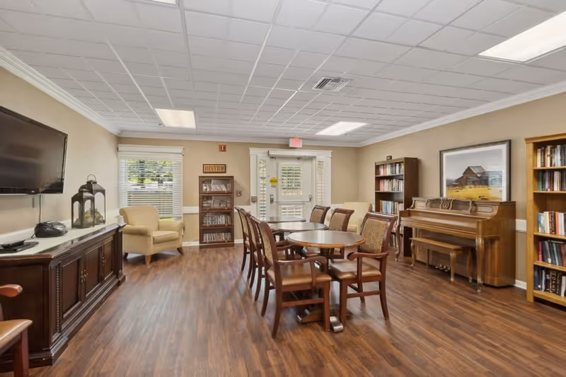 A well-lit common room with wooden flooring featuring a round table surrounded by chairs in the center. To the right, there is a wooden piano with a bench and bookshelves filled with books. On the left side, a flat-screen TV is mounted on the wall above a wooden cabinet, and a beige armchair is placed near a window. The room has a beige wall color and a white tiled ceiling with fluorescent lights. A door with glass panels is visible at the far end of the room.