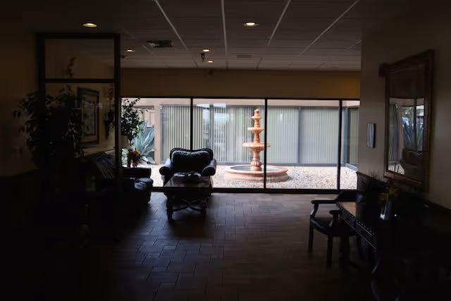 A dimly lit indoor seating area with armchairs and a coffee table facing a large window. Outside the window, there is a courtyard with a multi-tiered water fountain surrounded by gravel and plants.