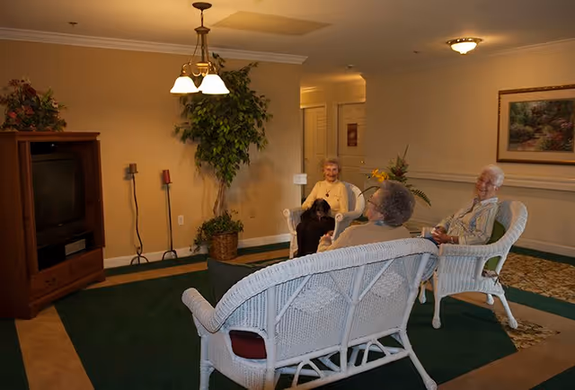 A cozy living room area in a senior living facility with three elderly women sitting on white wicker chairs and a loveseat, engaging in conversation. The room features a green carpet, a wooden TV stand with a television, a large potted plant, and a framed painting on the wall.
