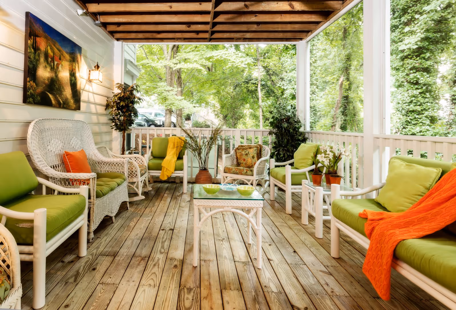 A cozy covered porch with wooden flooring and white railing, furnished with white wicker and wooden chairs and sofas with green cushions and colorful throw blankets. There are potted plants and a painting on the wall, with a view of lush green trees outside.