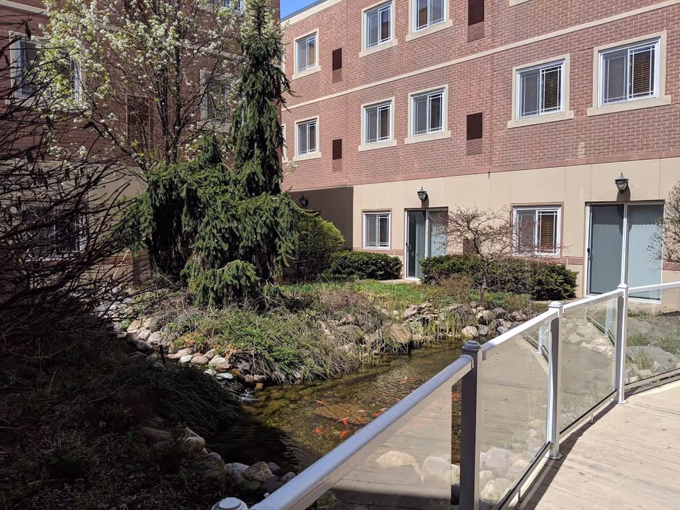 Outdoor courtyard area at Franciscan Court Supportive Living featuring a small pond with fish, surrounded by rocks and greenery including trees and bushes. The courtyard is enclosed by a brick building with multiple windows and doors, and a glass railing along a wooden walkway.