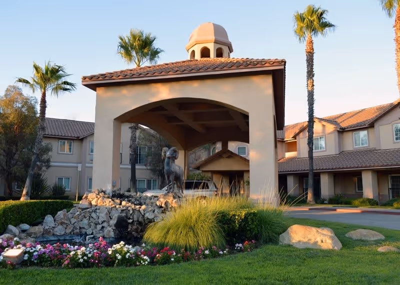 Entrance area of The Lakes Senior Living facility featuring a covered driveway with a tiled roof, a decorative ram statue on a rock formation surrounded by greenery and flowers, palm trees, and a two-story building in the background.