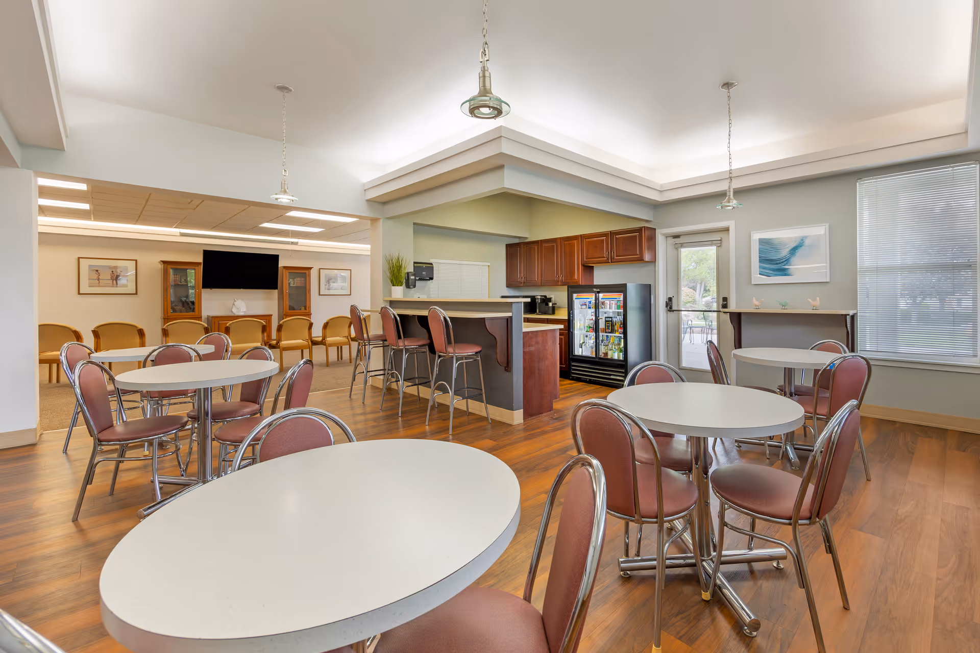 A bright and clean common area in a senior living facility featuring several round tables with pink cushioned chairs, a small bar area with high stools, a refrigerator stocked with beverages, and a seating area with chairs facing a wall-mounted TV. Large windows and a glass door provide natural light and a view of the outdoor patio.