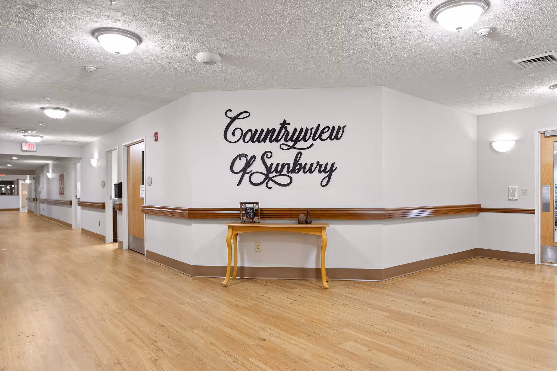 Interior hallway of a senior living facility with light wood flooring, white walls, and ceiling lights. A wooden table is placed against the wall with decorative items on it. The wall features a large decorative sign that reads 'Countryview of Sunbury'. Several doors line the hallway, and there is an exit sign visible in the distance.