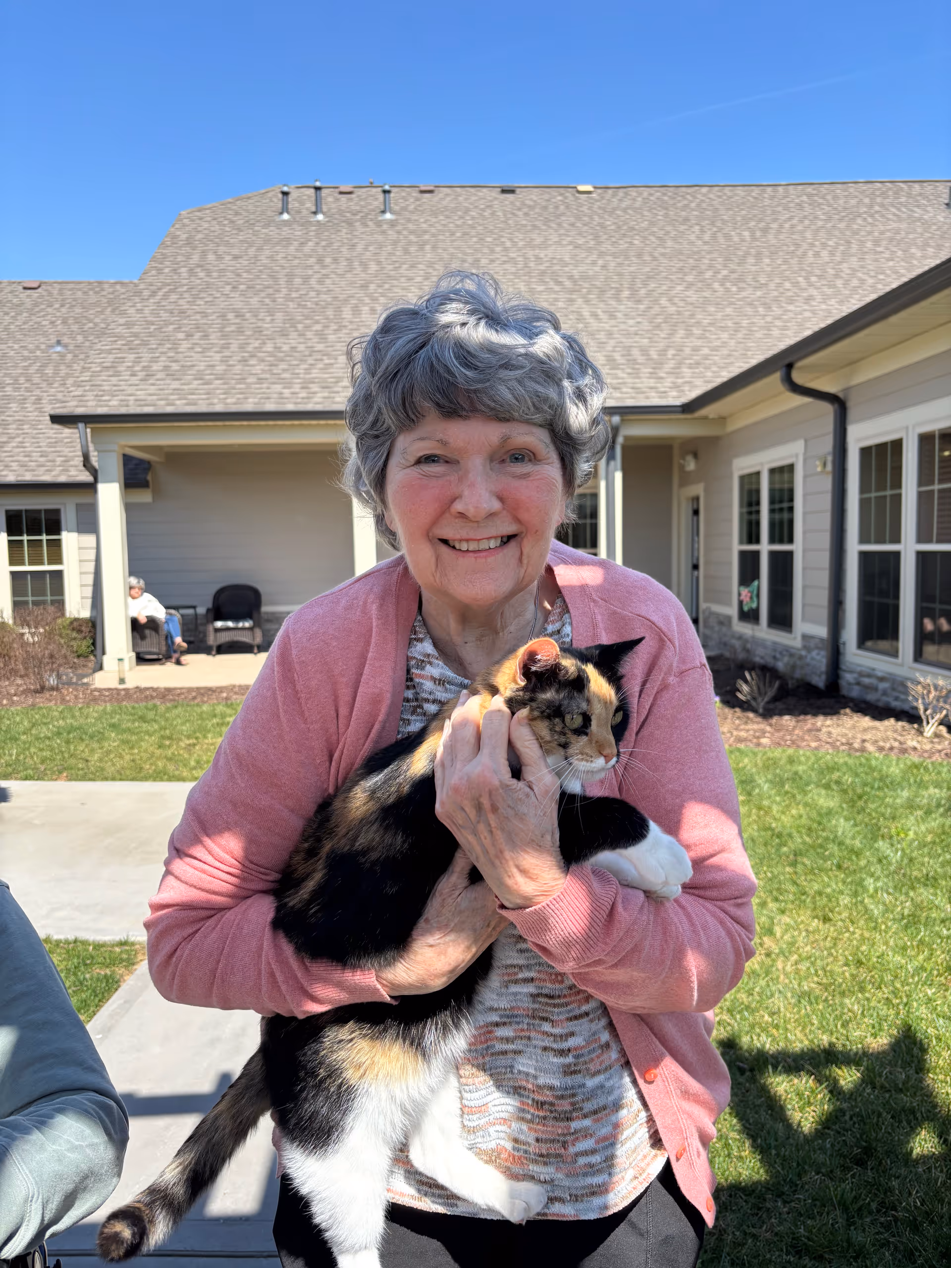 An elderly woman with gray hair wearing a pink cardigan and patterned shirt is smiling and holding a calico cat outside in a sunny courtyard area with grass and a building in the background.