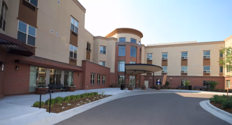 Exterior view of a multi-story senior living facility building with a curved driveway and entrance canopy. The building features a combination of brick and beige siding with multiple windows and some landscaping along the walkway.