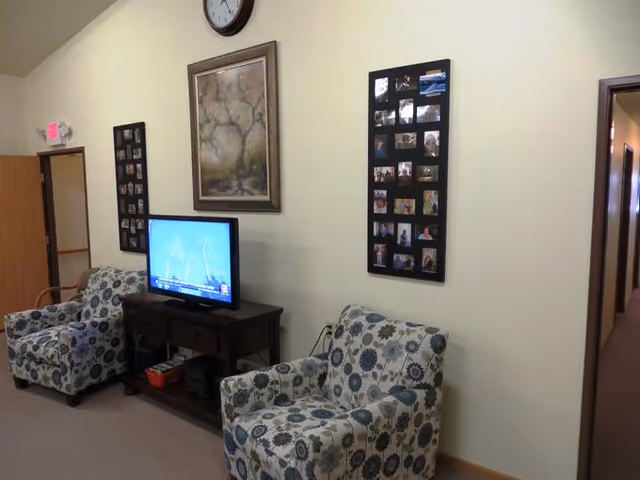 A cozy living area with two patterned armchairs facing a small wooden TV stand holding a flat-screen television. The wall behind the TV features a large framed painting of a tree and two photo collage frames. A clock is mounted above the painting. There is an open doorway on the left and a hallway on the right.