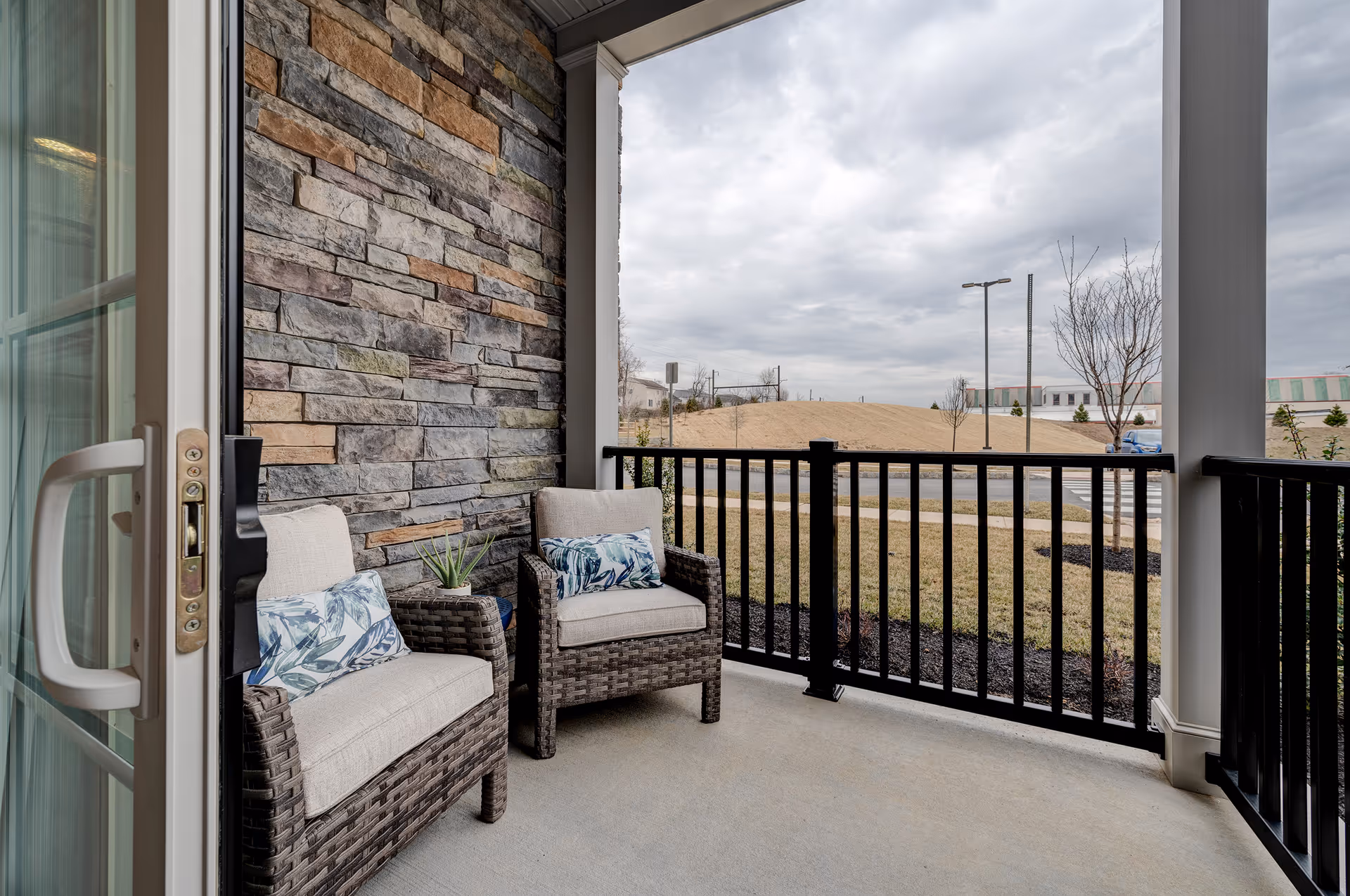 A covered outdoor patio area with two cushioned wicker chairs, each with a decorative pillow. The patio has a stone wall on one side and black railing overlooking a grassy area and a road with a cloudy sky in the background.