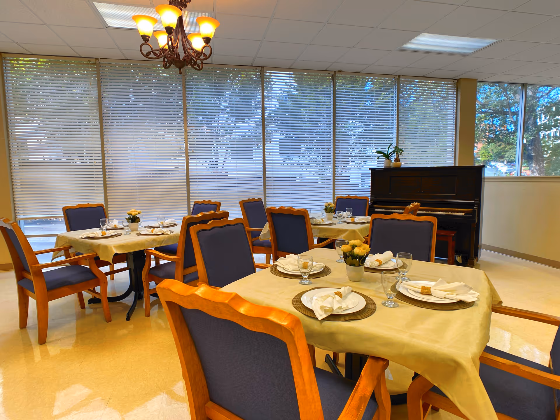 A dining room with several tables covered with beige tablecloths, each set with plates, napkins, glasses, and small flower arrangements. The room has large windows with blinds, a chandelier hanging from the ceiling, and a piano in the corner with a plant on top.