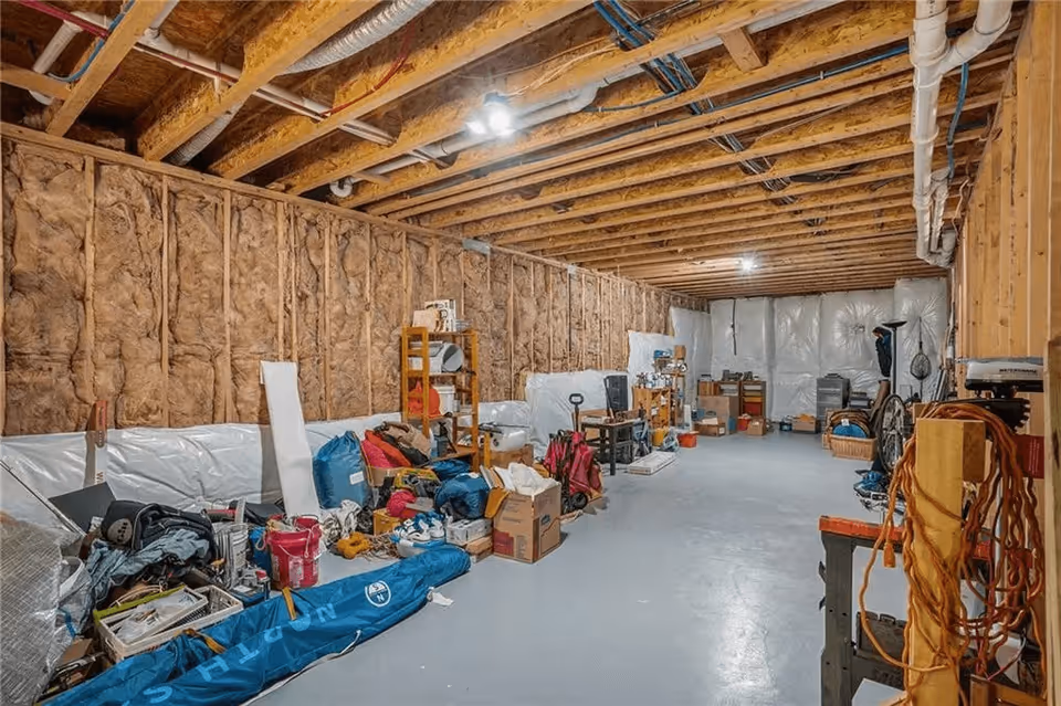 A large unfinished basement with exposed wooden beams and insulation on the walls. The floor is concrete and various household items, boxes, and equipment are scattered and stored along the walls. The ceiling has visible pipes and ductwork with overhead lighting.