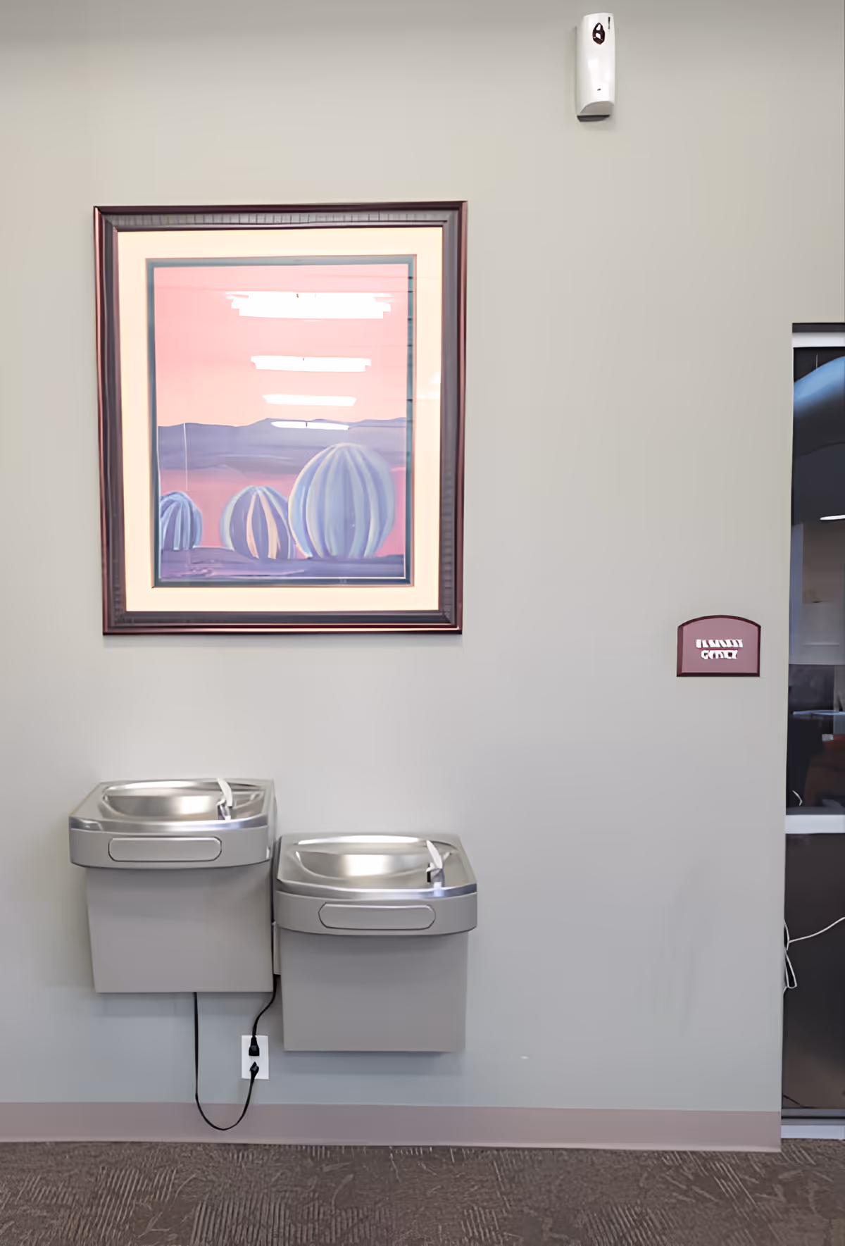 Two wall-mounted drinking fountains of different heights installed side by side on a gray wall. Above the fountains is a framed painting depicting abstract blue and purple shapes against a pink and purple background. To the right, there is a small maroon sign with white text, and part of a glass door or window is visible.