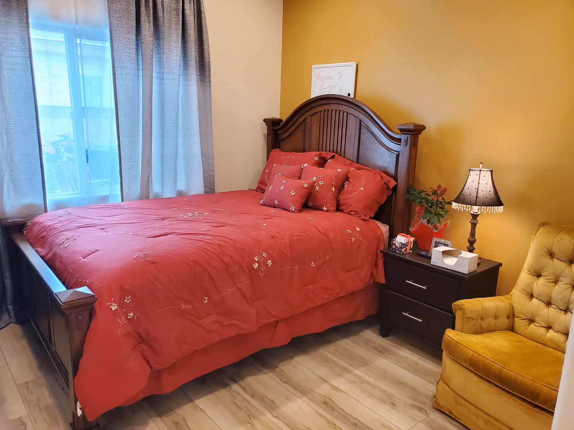 Cozy bedroom with a wooden bed dressed in red bedding, a nightstand with lamp and plant, and a yellow armchair.