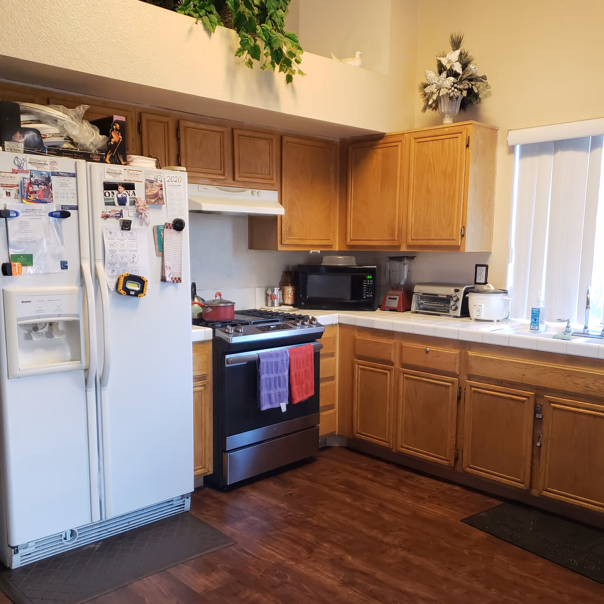 A kitchen with wooden cabinets, a white refrigerator covered with magnets and papers, a stainless steel stove with two towels hanging on the handle, a microwave, a blender, a toaster oven, and a rice cooker on the tiled countertops. There is a window with vertical blinds above the sink and decorative plants on top of the cabinets.