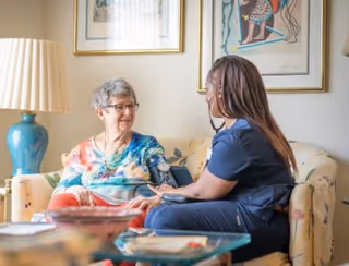 An elderly woman sitting on a floral-patterned couch smiling and holding hands with a healthcare worker dressed in navy scrubs, who is sitting next to her and taking her blood pressure with a cuff. The room has framed artwork on the wall and a blue lamp on a side table.