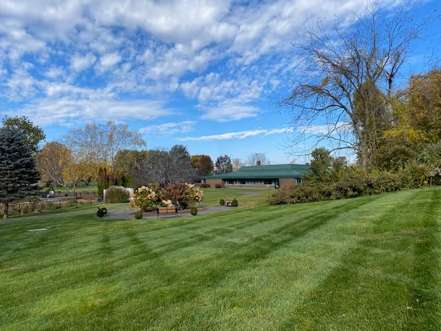 A large, well-maintained grassy lawn with a few benches and landscaped bushes in the foreground. In the background, there is a low building with a green roof surrounded by trees with autumn foliage under a partly cloudy blue sky.