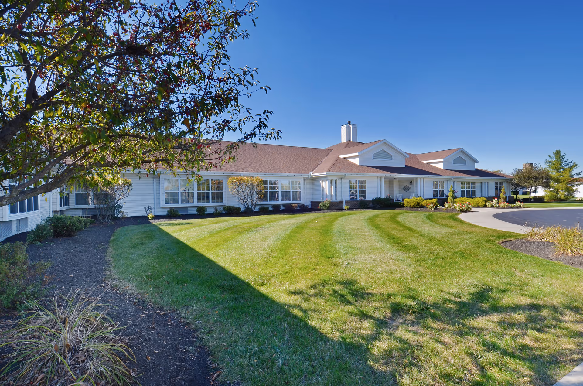 Exterior view of a single-story senior living facility building with white siding and a brown roof, surrounded by a well-maintained lawn and landscaping under a clear blue sky.