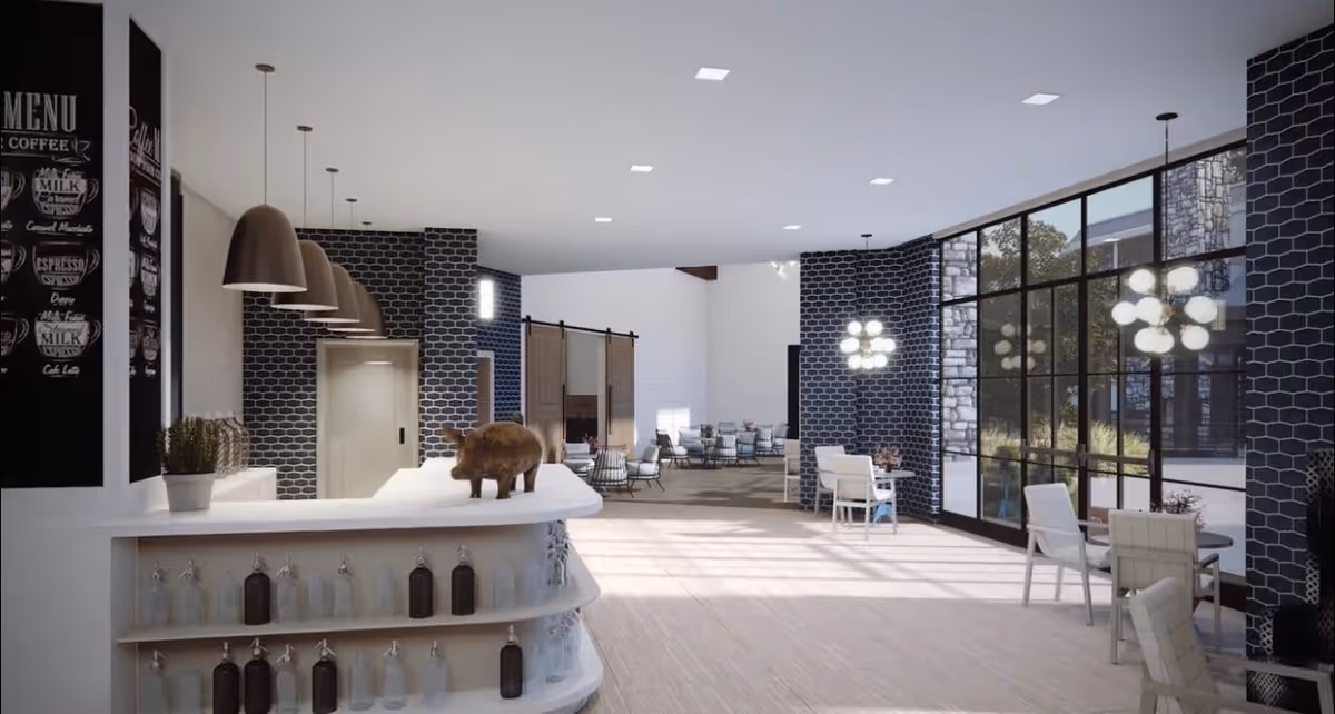 A bright and modern common area with large floor-to-ceiling windows letting in natural light. The space features a white curved counter with glass bottles on shelves underneath, pendant lights hanging above, and a decorative pig figurine on the counter. There are tables and chairs arranged near the windows and a seating area with armchairs in the background. The walls have a dark hexagonal tile pattern and white brick accents.