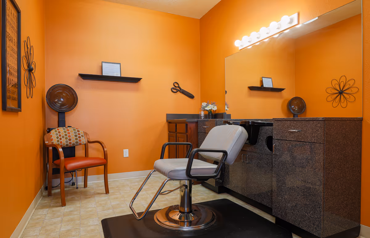 A brightly lit hair salon room with orange walls featuring a salon chair in front of a large mirror and countertop with cabinets. There is a hair dryer chair in the corner, a wooden chair with a patterned cushion, and decorative wall art including a large pair of scissors and flower shapes.