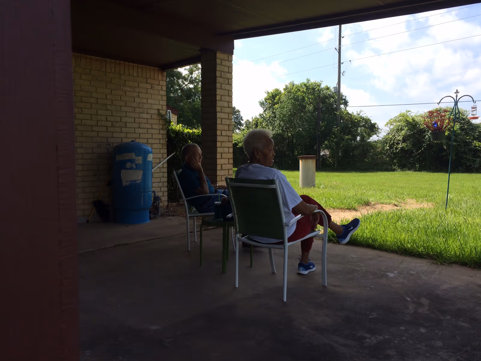 Two elderly people sitting on chairs under a covered patio, facing a grassy yard with trees and a blue sky in the background.