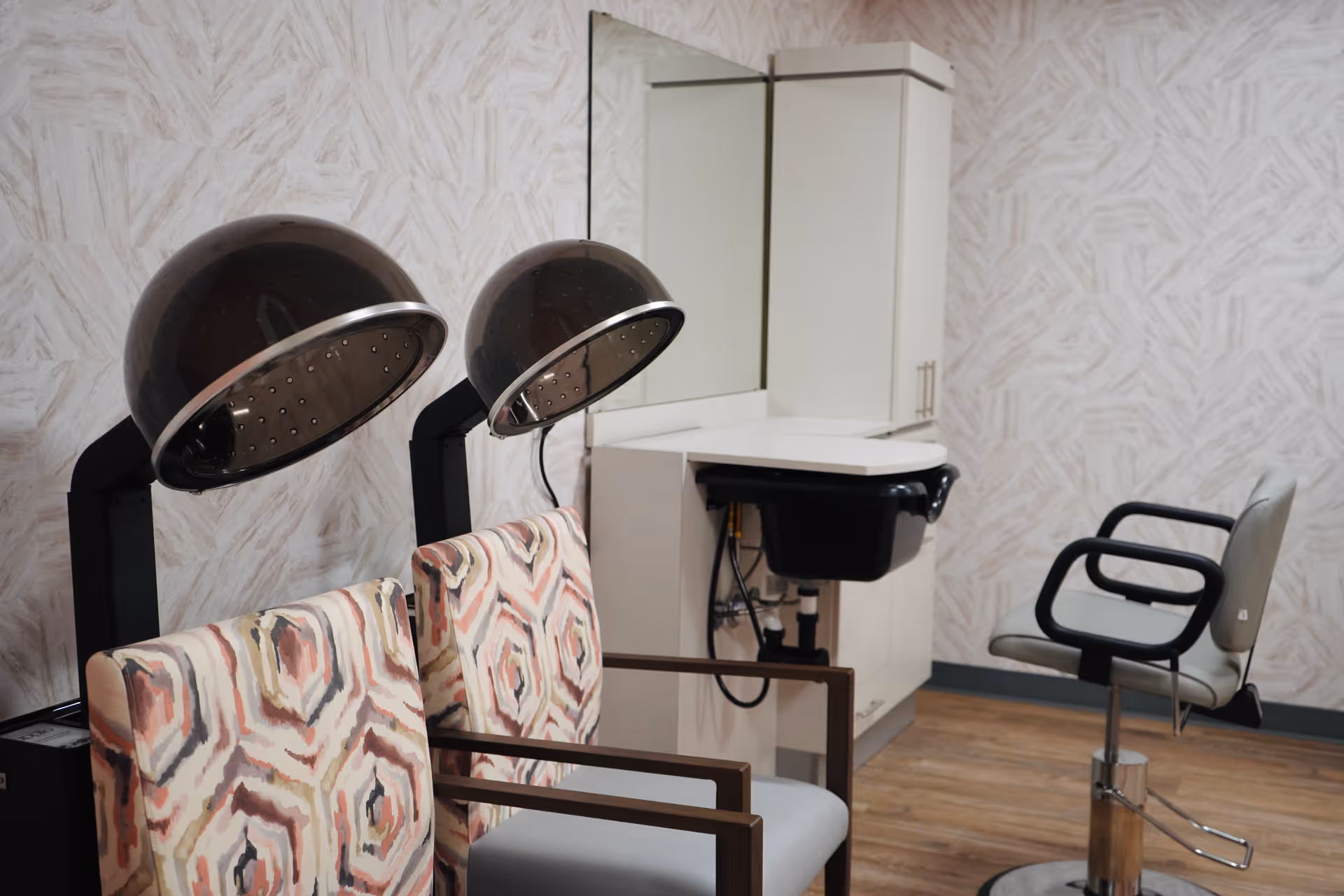 A small salon area with two hooded hair dryers, patterned waiting chairs, a styling sink and a salon chair against patterned wallpaper.