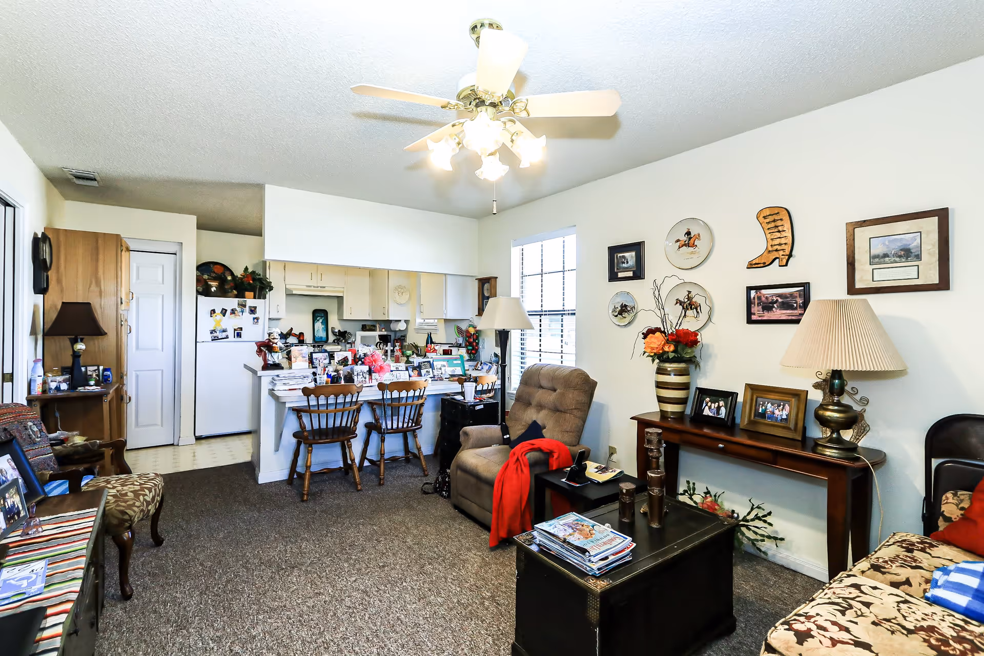 A cozy living room and kitchen area in a senior living facility. The living room features a cushioned armchair with a red blanket, a wooden coffee table with magazines, a side table with a lamp, and a console table with framed photos and a vase of flowers. The wall is decorated with framed pictures and decorative plates. The kitchen has white cabinets, a refrigerator, and a counter with two wooden chairs. The room is well-lit with a ceiling fan light and natural light from a window.