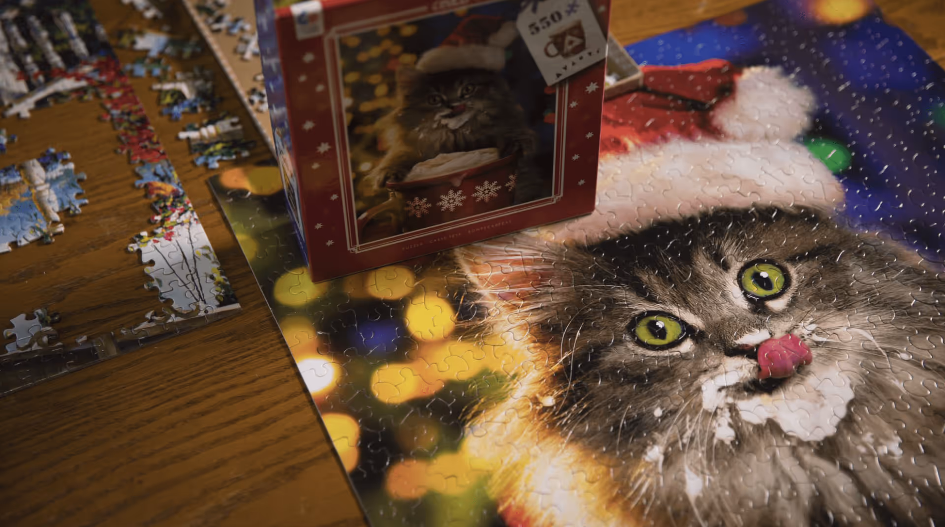 A partially completed jigsaw puzzle on a wooden table featuring a close-up image of a cat wearing a Santa hat with its tongue out and some white cream around its mouth. The puzzle box with the same cat image is placed on top of the puzzle.