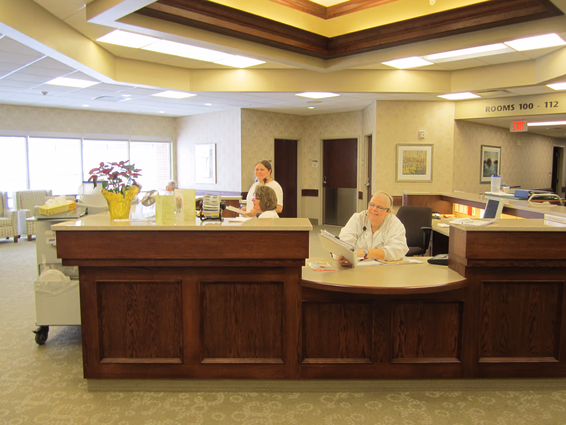 Reception area in a nursing and rehab center with a wooden front desk. Three staff members are behind the desk, one sitting and smiling while holding a folder, and two others standing and talking. The area is well-lit with large windows and comfortable seating visible in the background. A sign above the hallway indicates rooms 100 to 112.