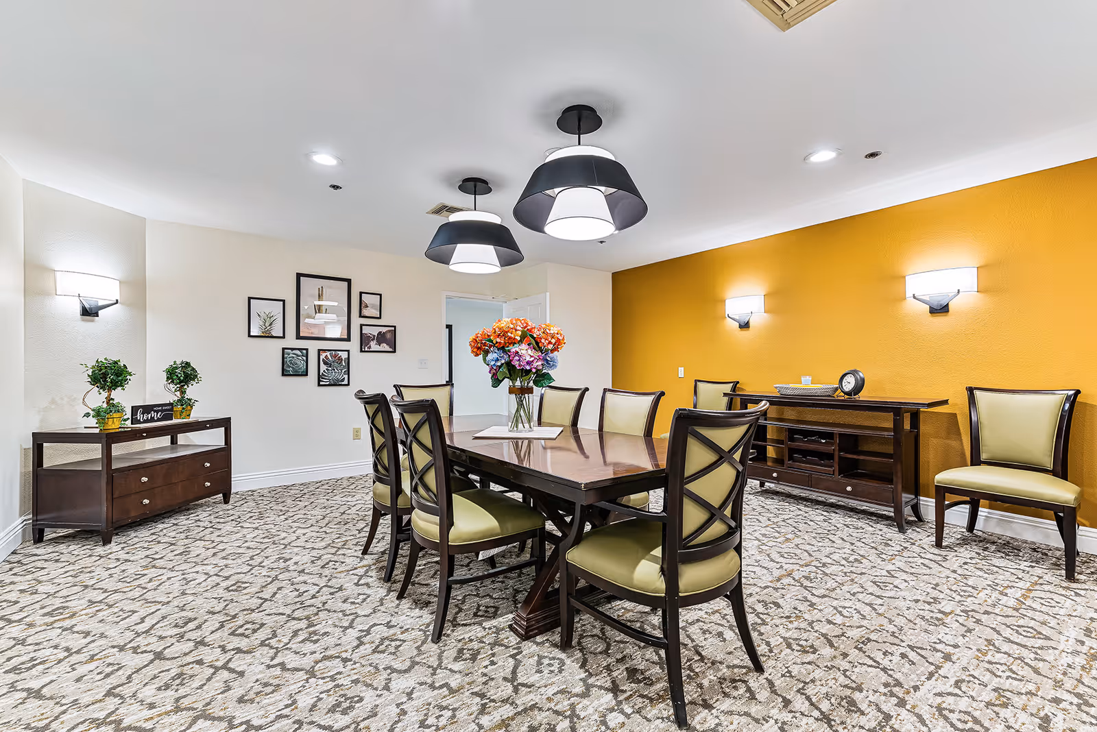 A dining room with a rectangular wooden table surrounded by eight chairs with green cushions. The room features a patterned carpet, a yellow accent wall with two wall sconces, and two modern pendant lights hanging from the ceiling. There are framed pictures on one wall and two wooden sideboards, one with plants and a decorative sign, and the other with a clock and a bowl.